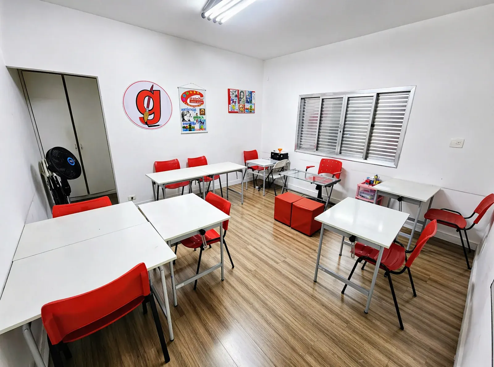 Sala de Artes visuais com mesas brancas, cadeiras vermelhas e logo Galpão na parede